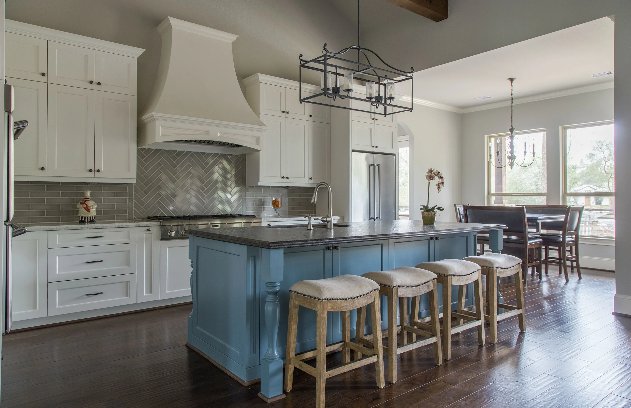 a modern texas kitchen undergoing a stylish remodel showcases a sleek, open layout with advanced 3d renderings displayed on a designer's tablet, surrounded by elegant cabinetry samples, vibrant materials, and a team of focused professionals engaged in a detailed planning discussion. Stylish kitchen with white cabinetry, a striking blue island topped with dark countertop, four cushioned bar stools, herringbone backsplash, and modern pendant lighting, opening into a dining area with large windows.