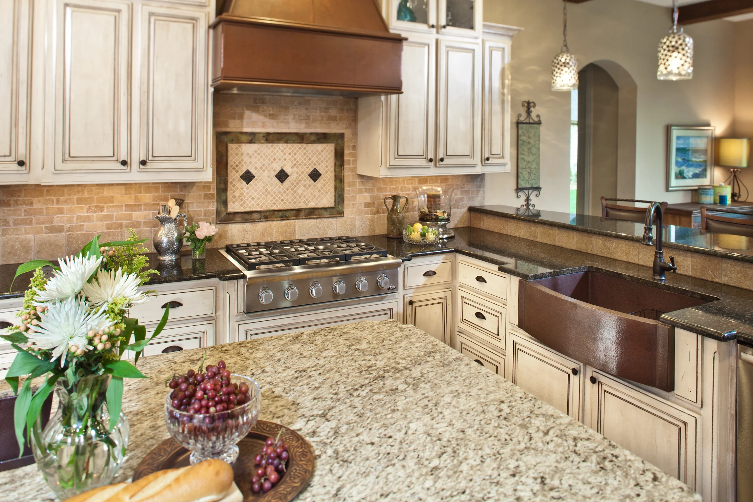 an elegantly designed luxury kitchen showcases stunning granite countertops, custom hardwood cabinetry, and sleek porcelain tile flooring, all illuminated by soft, ambient lighting that highlights the rich textures and finishes. Warm, rustic-style kitchen featuring cream-colored distressed cabinetry, a granite countertop island, copper farmhouse sink, decorative tile backsplash, and pendant lighting, accented with flowers, bread, and grapes on the counter.