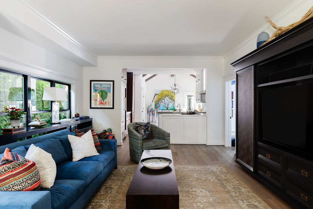 Bright living room with European White Oak flooring, a deep blue sofa with colorful pillows, large windows with black framing, and a view into the kitchen and dining area where Sherwin Williams Snowbound-painted walls and modern décor are visible.